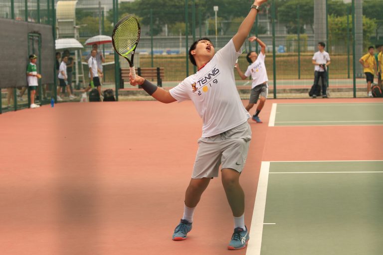 An ACS(I) tennis player stretches wide to return a shot during a competitive match on a red clay-style court.